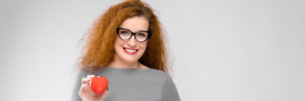 Encantadora mujer joven con el pelo rojo. Una mujer joven con un suéter gris. Una mujer joven con gafas está sosteniendo un corazón rojo