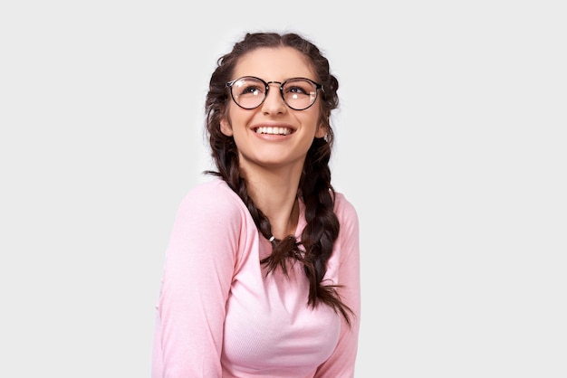 Encantadora mujer joven que sonríe ampliamente viste una blusa casual rosa y gafas transparentes redondas mirando hacia arriba aisladas sobre fondo blanco Mujer joven feliz con peinado de trenzas en la pared gris del estudio