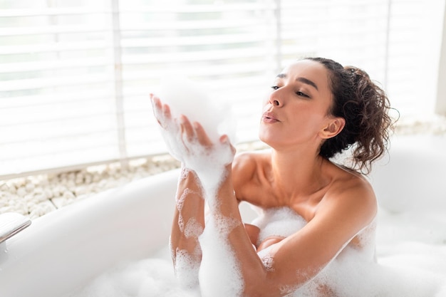 Encantadora mujer joven que sopla burbujas de jabón mientras se relaja en el baño en el espacio libre de casa