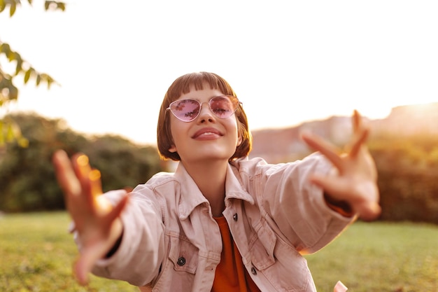 Foto encantadora mujer joven quiere abrazos y se estira a la cámara chica feliz en chaqueta de mezclilla rosa y elegantes gafas de sol camina al aire libre en el parque