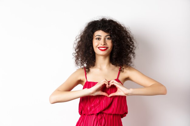 Foto encantadora mujer joven con rojo romántico fress, mostrando gesto de corazón y sonriendo, expresa amor, de pie sobre fondo blanco.