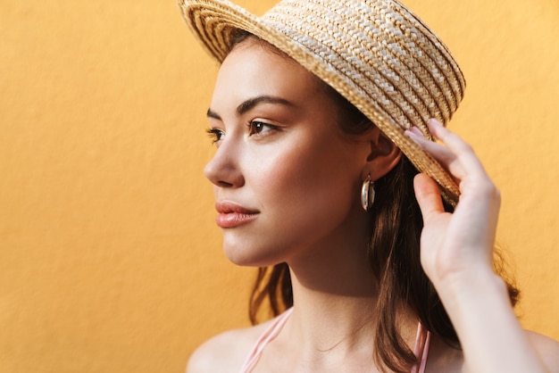 Encantadora mujer joven con sombrero de paja de verano mirando a un lado aislado en amarillo