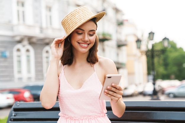 Encantadora mujer joven con sombrero de paja de verano sonriendo y sosteniendo el teléfono celular mientras camina por las calles de la ciudad