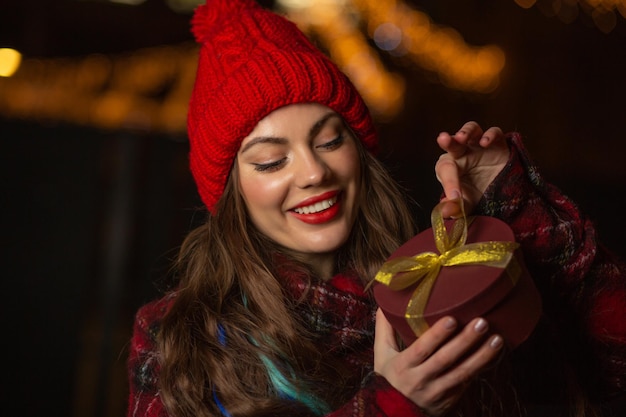 Encantadora mujer joven con sombrero rojo recibiendo un regalo en una caja en la feria de Navidad por la noche. Espacio vacio