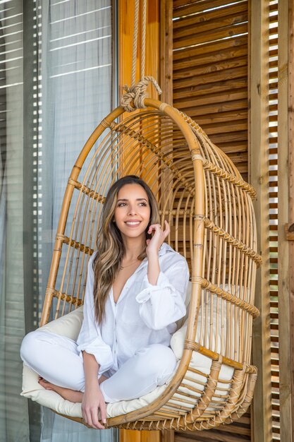 Encantadora mujer joven sonriente en ropa blanca sentada en una cómoda silla colgante de mimbre de ratán mientras se relaja en la terraza de la casa