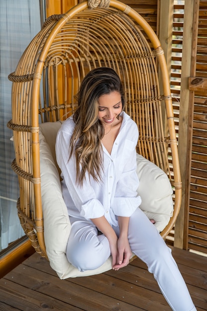 Encantadora mujer joven sonriente en ropa blanca sentada en una cómoda silla colgante de mimbre de ratán mientras se relaja en la terraza de la casa