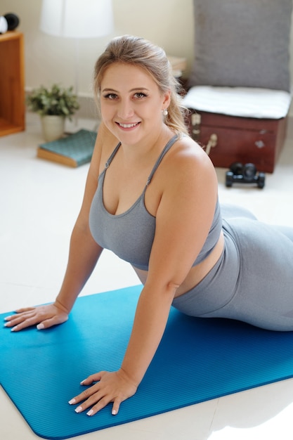 Encantadora mujer joven y sonriente de talla grande haciendo cobra asana al practicar yoga en casa