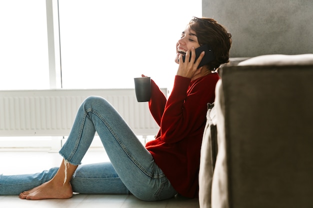 Encantadora mujer joven con suéter sentado en el suelo en casa, bebiendo té, apoyado en un sofá