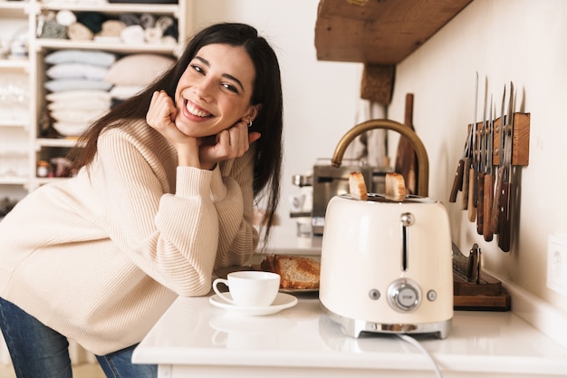 Encantadora mujer joven tomando una taza de café en la cocina, haciendo tostadas