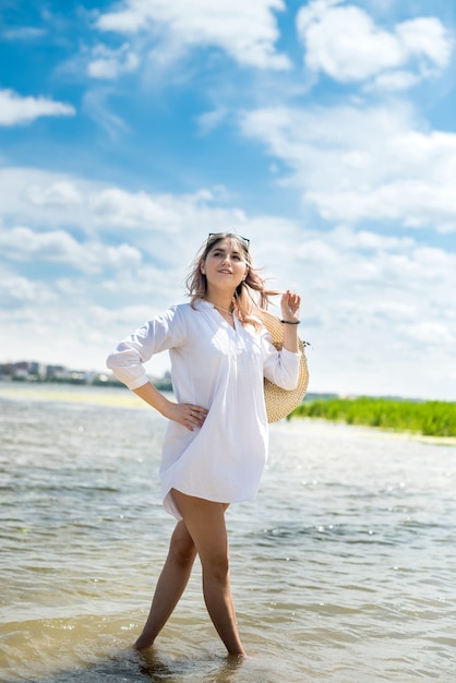 Encantadora mujer joven con un vestido blanco disfrutar de la naturaleza junto al lago en un día soleado. Sesión de fotos de moda con estilo