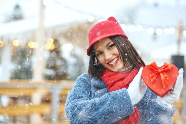 Encantadora mujer morena en abrigo de invierno sosteniendo una caja de regalo en la feria de Navidad durante las nevadas. Espacio para texto