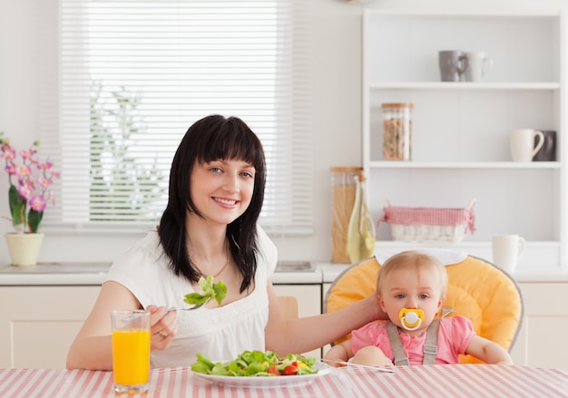 Encantadora mujer morena comiendo una ensalada al lado de su bebé mientras está sentado