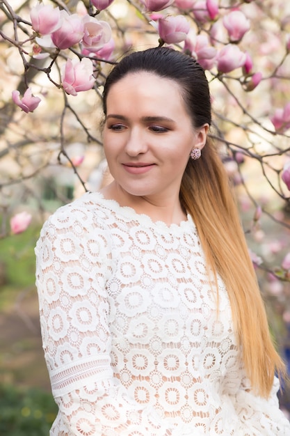 Encantadora mujer morena con maquillaje nude, vistiendo blusa de encaje, posando cerca de las flores de magnolia en flor