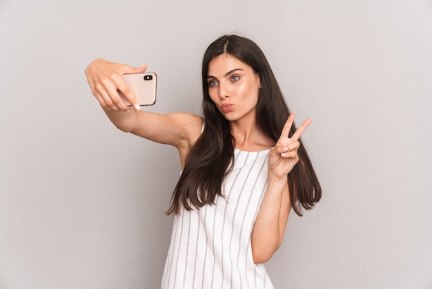 Encantadora mujer morena con un vestido que muestra el signo de la paz mientras toma una foto selfie en un teléfono inteligente aislado sobre una pared gris