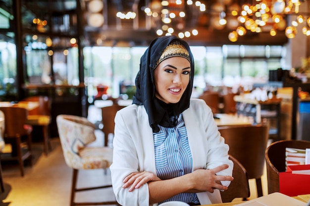 Encantadora mujer musulmana sonriente moderna con bufanda sentada en la cafetería con las manos cruzadas