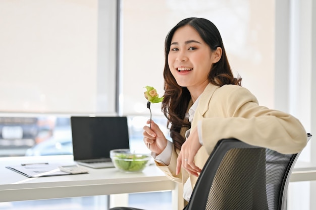 Encantadora mujer de negocios asiática está en su escritorio con una ensaladera saludable almorzando