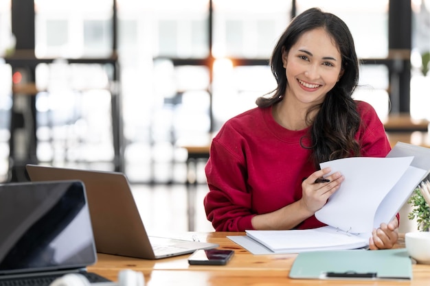 Encantadora mujer de negocios asiática que trabaja con papeleo y computadora portátil para analizar datos comerciales y financieros en la oficina sonriendo y mirando la cámara