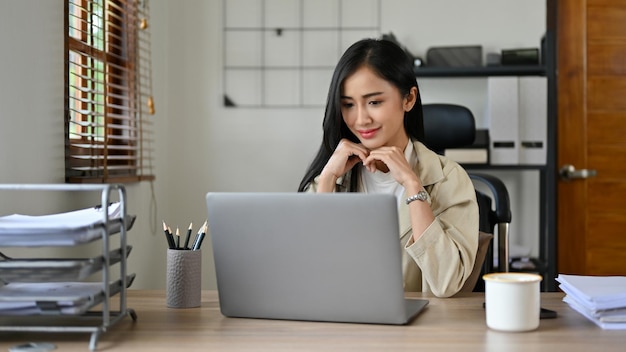 Encantadora mujer de negocios asiática se sienta en su escritorio mirando la pantalla de su computadora portátil