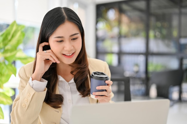 Encantadora mujer de negocios asiática tomando café mientras trabaja en su proyecto en una laptop en la oficina