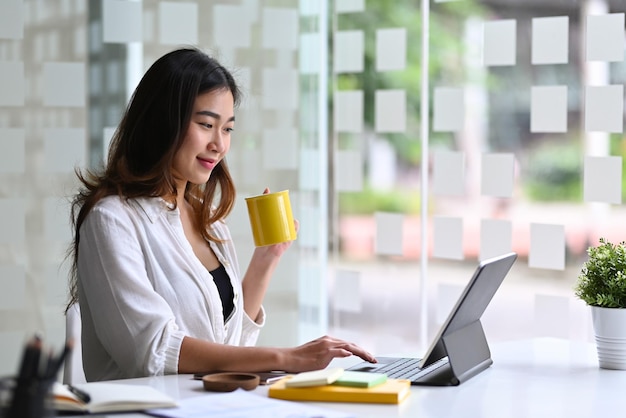 Encantadora mujer de negocios bebiendo café y revisando el correo electrónico en la tableta de la computadora