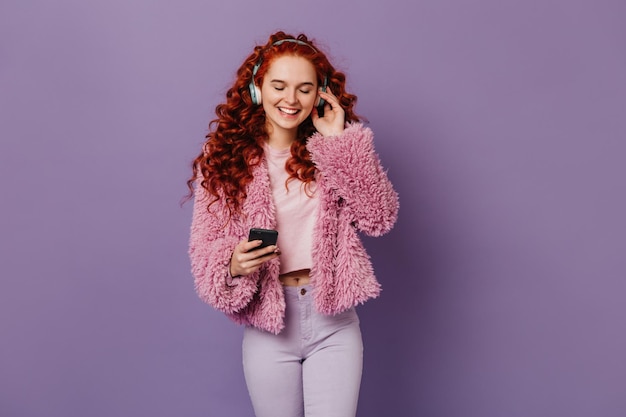 Encantadora mujer pelirroja con pantalones blancos, top rosa y abrigo, sonríe con auriculares y sostiene el teléfono