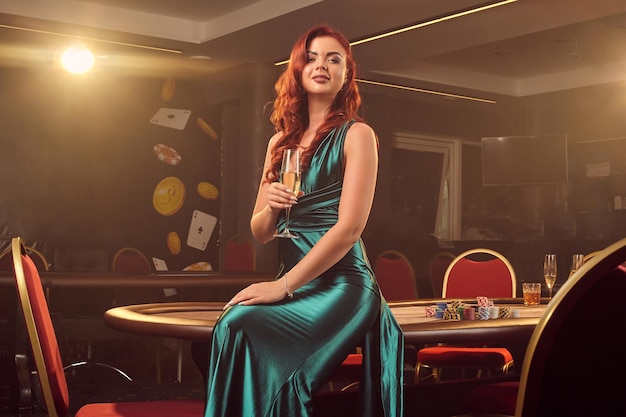 Encantadora mujer pelirroja con un vestido largo de satén azul, con una copa de champán en la mano, sonríe y posa sentada en una mesa de póquer en un casino de lujo. Pasión, cartas, fichas, alcohol, ganar, gambli