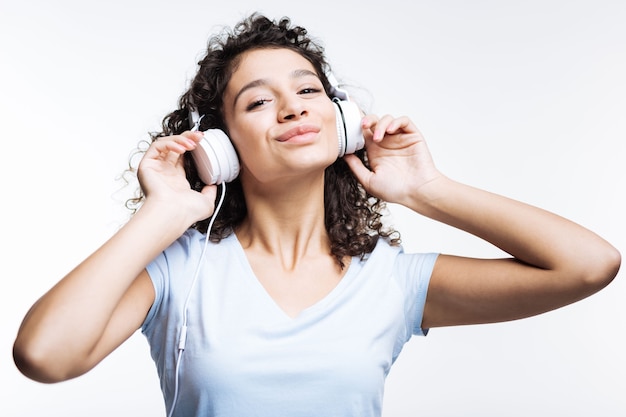 Encantadora mujer de pelo rizado en una camiseta blanca escuchando música en auriculares y sonriendo mientras está de pie aislado en gris