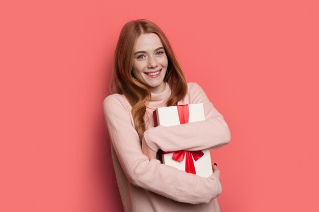 Encantadora mujer de pelo rojo con pecas sonriendo a la cámara mientras abraza un regalo en un cálido suéter posando en una pared roja