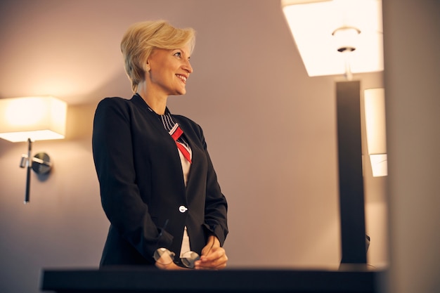 Foto encantadora mujer rubia en elegante chaqueta, mirando a otro lado y sonriendo mientras sostiene gafas