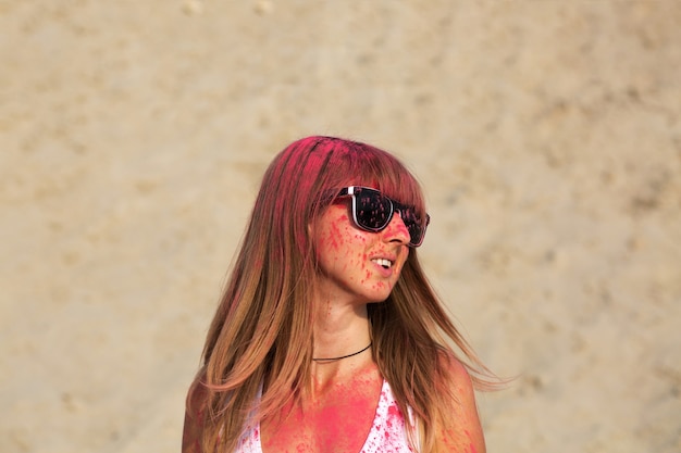 Encantadora mujer rubia con gafas jugando con pintura seca rosa Holi en el desierto