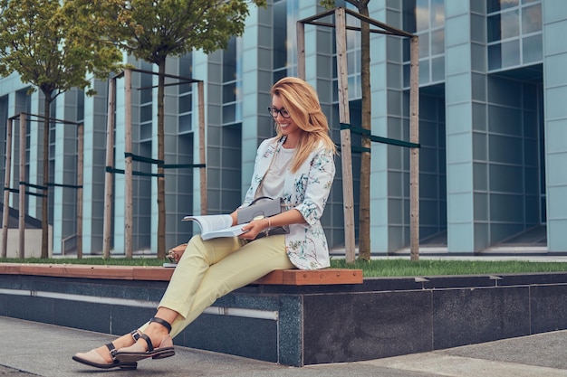 Encantadora mujer rubia con ropa moderna, estudiando con un libro, sentada en un banco en el parque contra un rascacielos