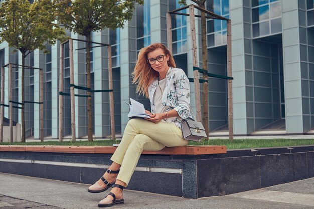Encantadora mujer rubia con ropa moderna, estudiando con un libro, sentada en un banco en el parque contra un rascacielos