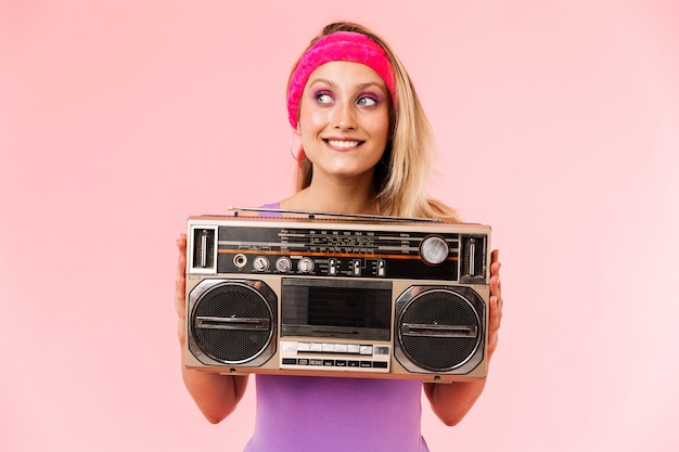 Encantadora mujer sonriente en diadema sosteniendo retro tape-player y mirando a un lado aislado sobre pared rosa