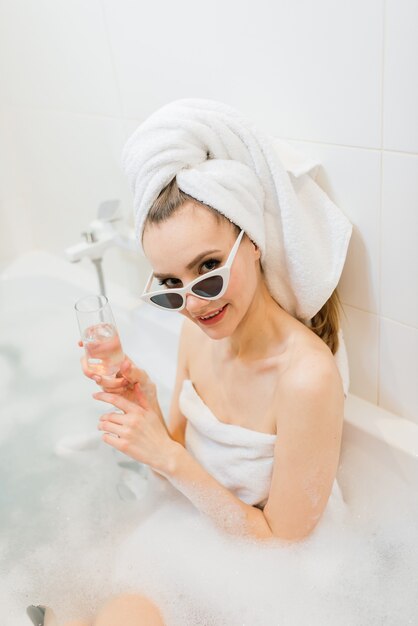 Encantadora mujer con sonrisa blanca como la nieve se sienta en el baño. Mujer elegante en toalla en la cabeza y gafas de sol toma tratamientos de spa en casa u hotel