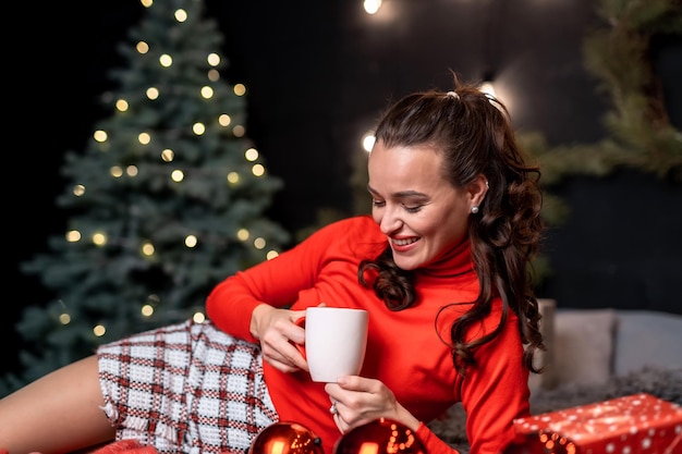 Encantadora mujer tendida en la cama bebiendo té y posando. Señora mirando taza. Foto de año nuevo.