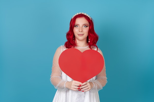 Encantadora mujer vestida de blanco y con cabello rojo sosteniendo un gran corazón de papel rojo