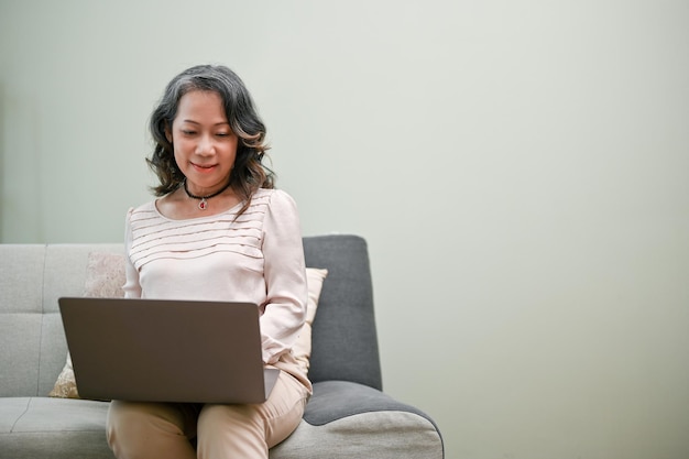 Encantadora mulher agedasiana usando laptop sentado em um sofá em sua sala de estar