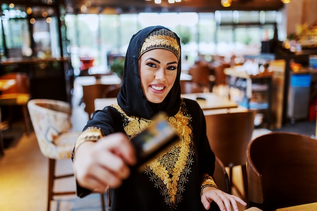Encantadora mulher árabe positiva com sorriso lindo vestido com roupas tradicionais, sentado no café