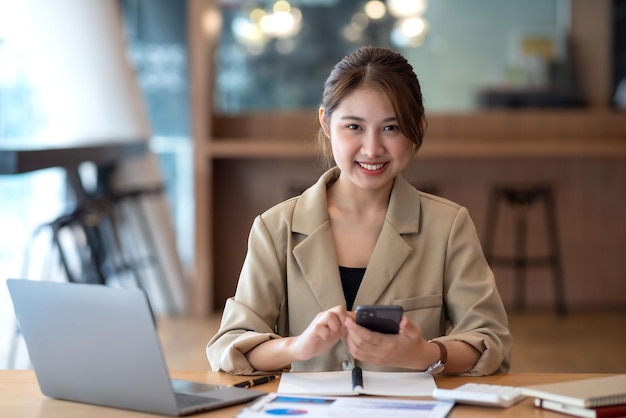 Encantadora mulher asiática com um sorriso sentado segurando smartphone no escritório