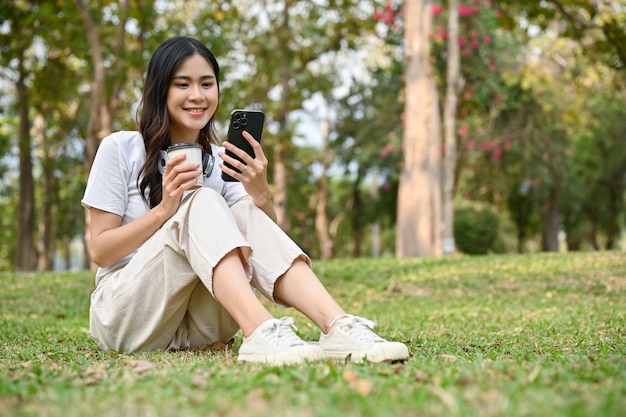 Encantadora mulher asiática senta-se na grama tomando café e usando seu telefone enquanto relaxa no parque