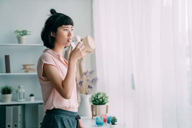 encantadora mulher chinesa asiática bebendo água na vista da janela no sol da manhã na sala de estar em casa. senhora feliz desfrutar de um copo de água fresca no apartamento moderno. relaxe a mulher aproveite a vista urbana com sol