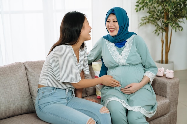 encantadora mulher chinesa asiática visitando amiga muçulmana grávida na sala de estar de casa. senhora alegre tocando a barriga grande da irmã de maternidade no sofá no apartamento. meninas felizes se divertindo no chá de bebê.