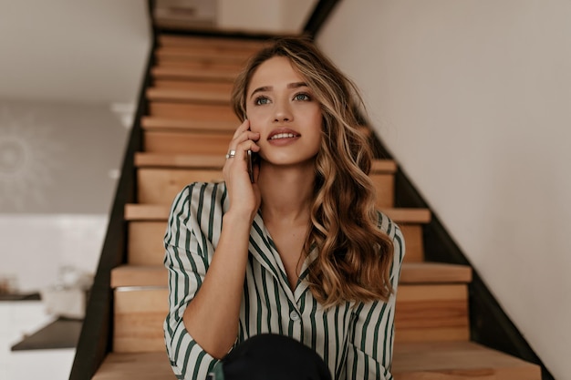 Encantadora mulher encaracolada de camisa listrada de seda verde e branca sentada em escadas de madeira fala no celular