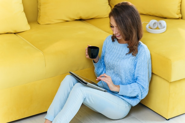 Encantadora mulher exuberante segurando seu ipad nas mãos, sentada no chão perto do sofá verde claro e desfrutando de café na xícara na cozinha contemporânea