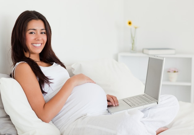 Encantadora mulher grávida relaxando com o laptop enquanto estava deitada em uma cama