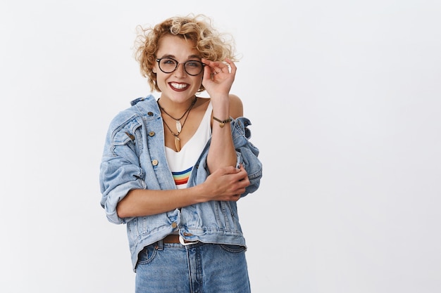 Foto encantadora mulher jovem, entusiasmada e feliz, rindo com um penteado curto loiro tocando as armações dos óculos olhando para a frente com alegria e rindo e se divertindo sobre a parede branca