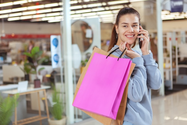 Encantadora mulher jovem falando ao telefone, andando com sacolas de compras no shopping