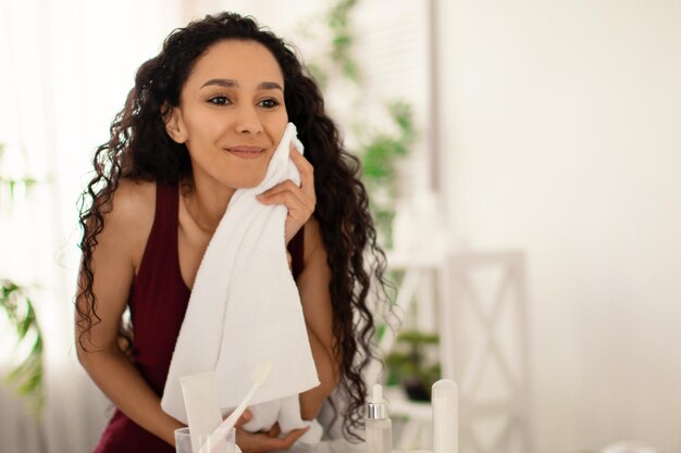 Encantadora mulher milenar realizando rotina de beleza matinal limpando o rosto com toalha olhando