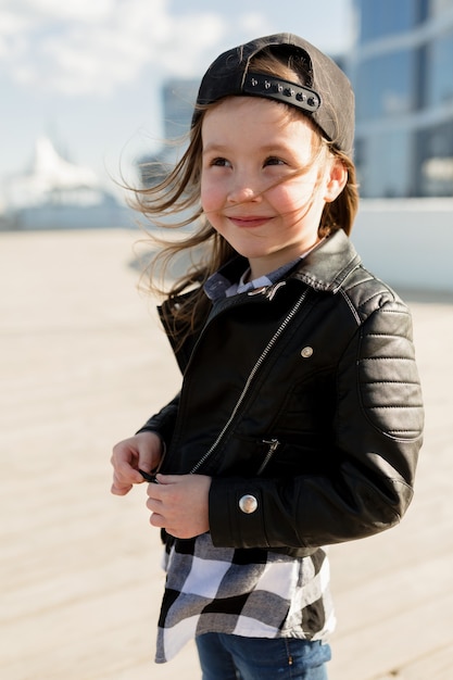 niña vestida con chaqueta de cuero y gorra sonriendo caminando cerca del mar en la luz del sol | Premium