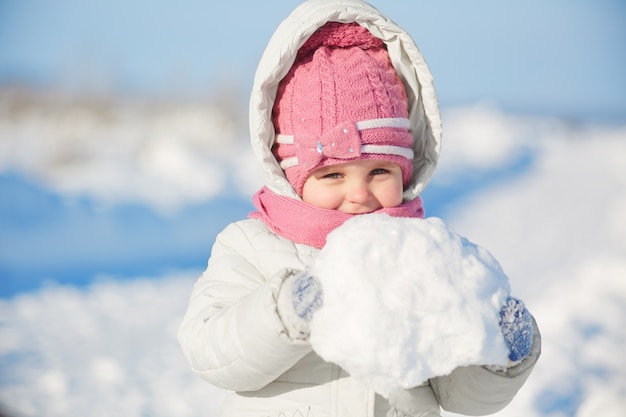 Encantadora niña bonita vestida con ropa de invierno cálida, sostiene bola de nieve se relaja al aire libre durante el clima helado del invierno, va a hacer un muñeco de nieve